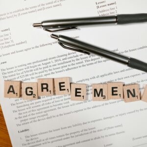 A document with Scrabble tiles spelling 'AGREEMENT' and two pens, symbolizing contract signing.
